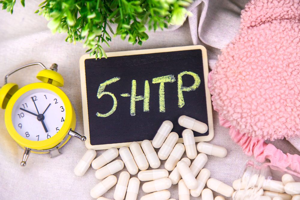 Egy asztalon fehér kapszulák, ébresztőóra és rózsaszín anyag veszi körül az „5-HTP” feliratú táblát.