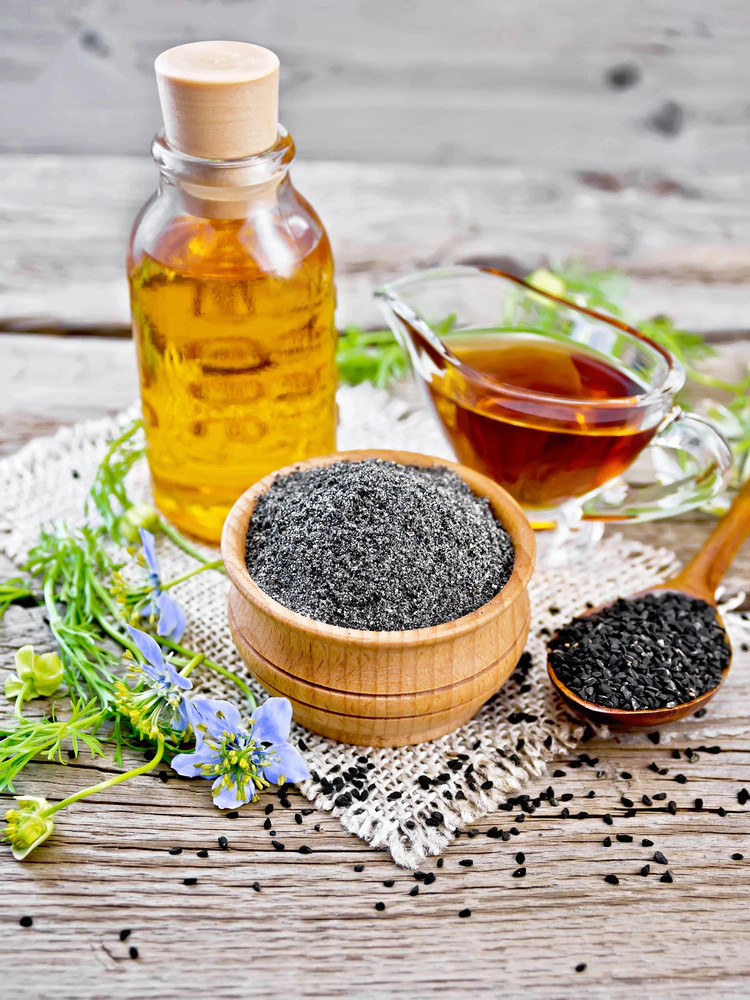 Fából készült tál fekete feketekömény magokkal, üvegek olajjal és Nigella Sativa növény rusztikus faasztalon.