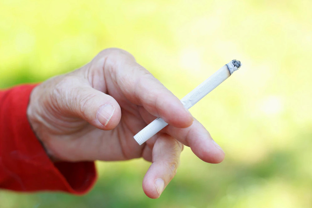 Kéz, amely hamus végű cigarettát tart; a háttér elmosódott, zöldes-sárgás árnyalatú.