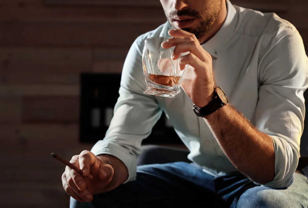 Fehér inges férfi whiskys poharat tart a kezében és szivart szív zárt térben – közelkép.
